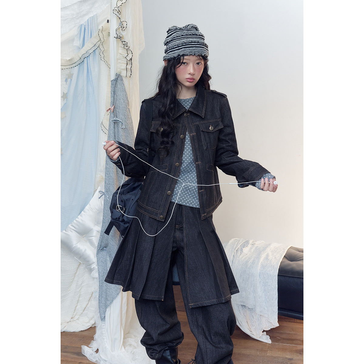 Model wearing a dark denim jacket, layered denim wrap skirt and wide-leg trousers with a striped knit cap.