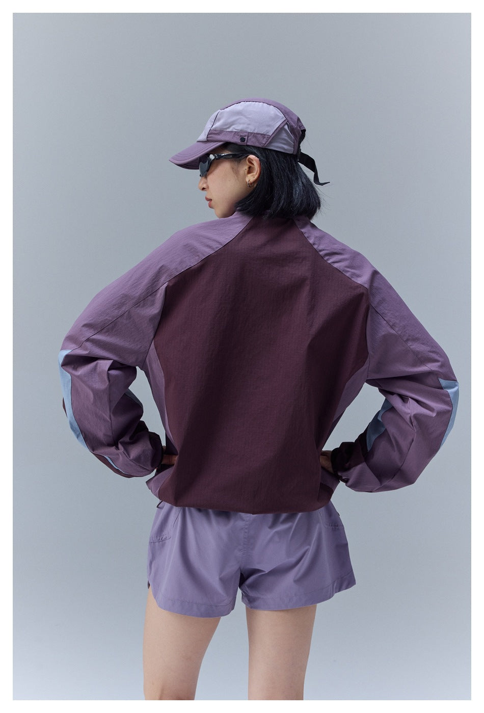 Back view of a model wearing the plum and purple color-block jacket with lavender shorts and a cap.