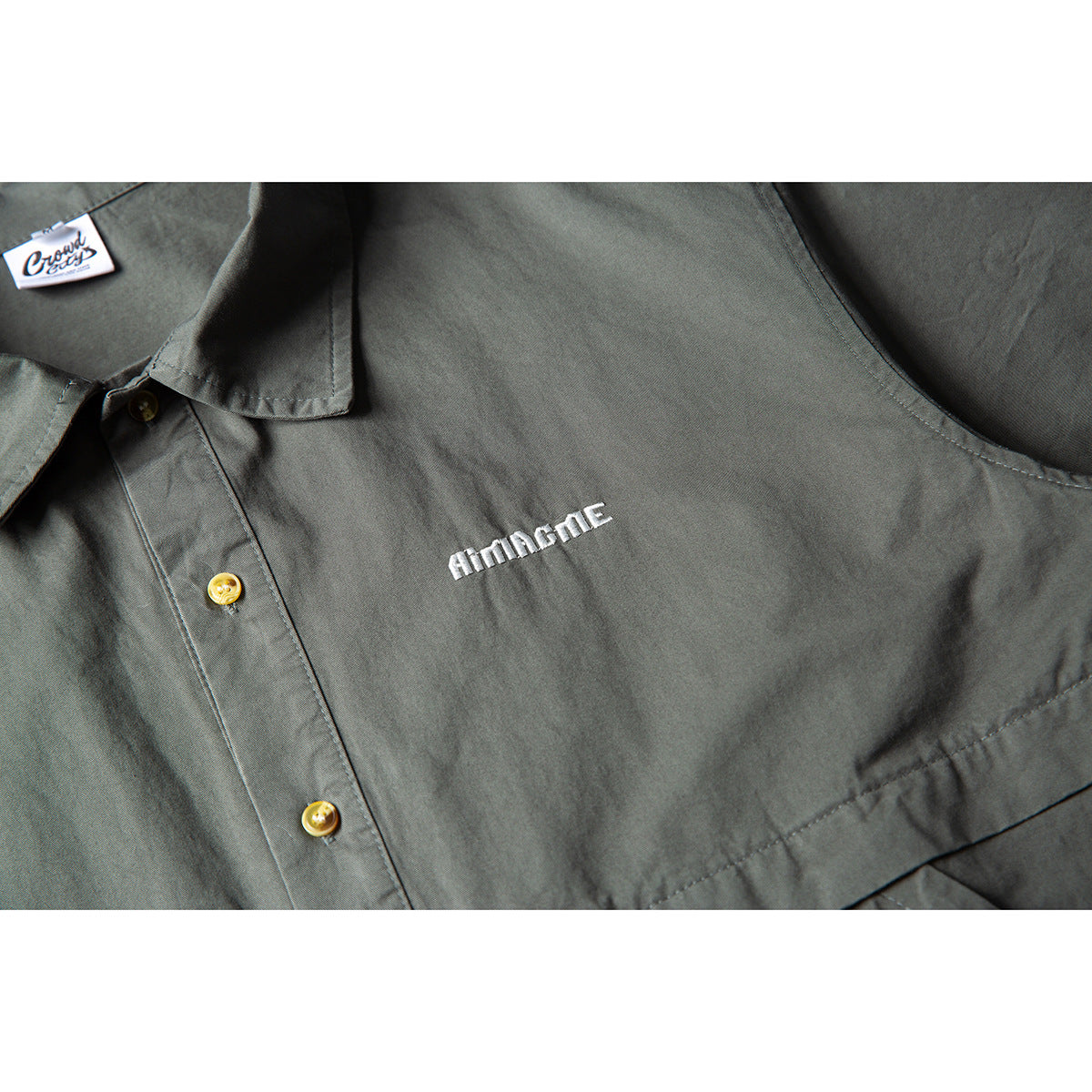 Close-up of gray-green shirt fabric, gold-toned buttons, and white embroidered chest lettering.