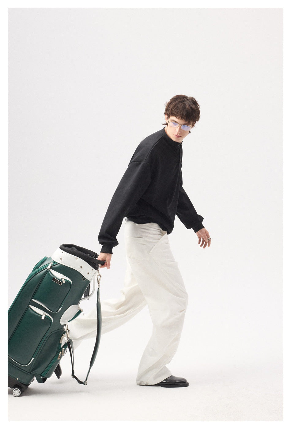 Model walking in a black button-neck top and white wide-leg trousers while pulling a green golf bag.