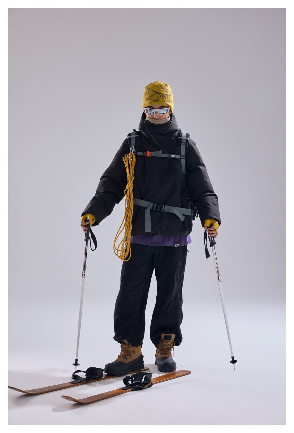 Front view of a model in a black hooded pullover and trousers, wearing a backpack and standing on skis.