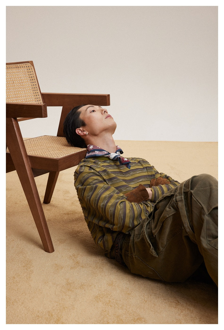 Model reclining in an olive and brown striped long-sleeve shirt, patterned neck scarf, and dark olive trousers.
