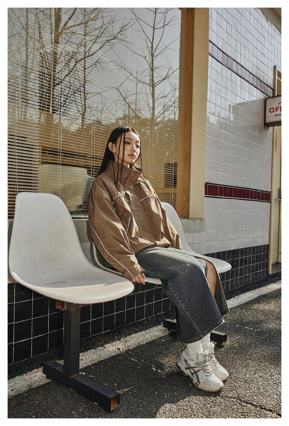 A seated model wears a brown jacket with white piping and green accents, a long denim skirt, and white sneakers.