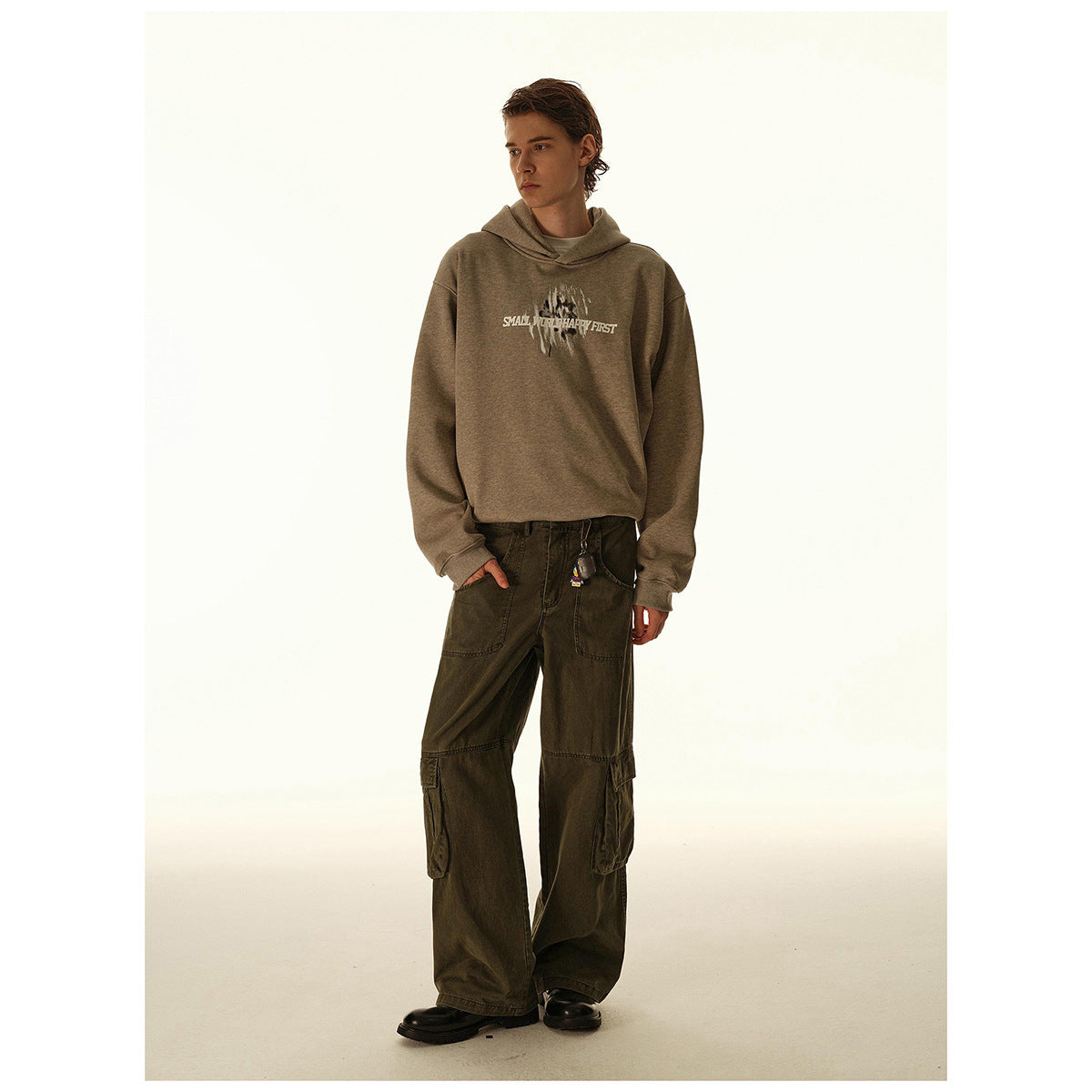 Full-length model wearing a taupe graphic hoodie, loose dark cargo pants, and black shoes.