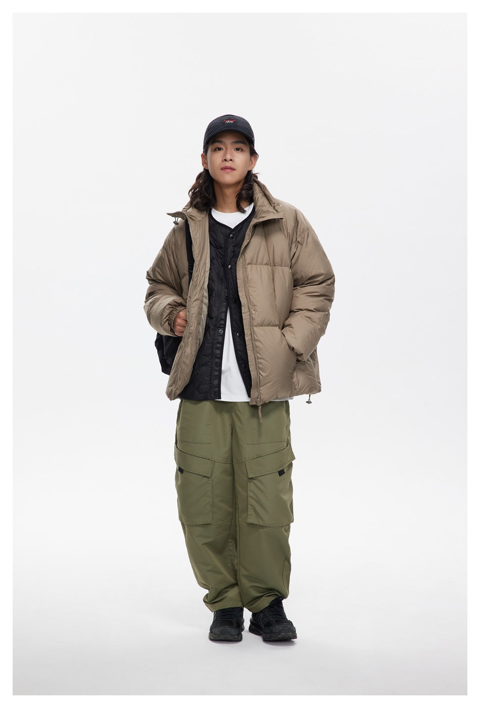 Model wearing a taupe puffer jacket open over a black vest and white shirt, with olive cargo pants.