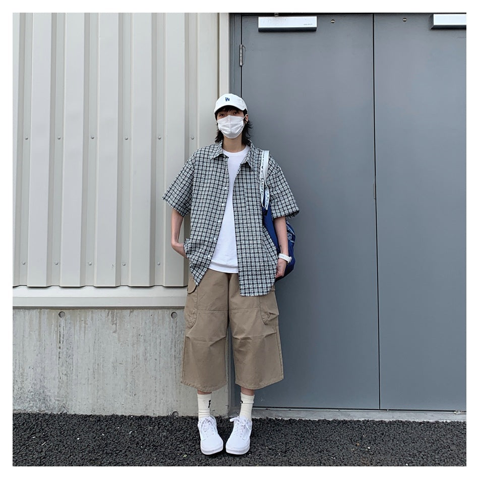 Person wearing an open black plaid short-sleeve shirt over a white tee with khaki cropped pants against a gray door