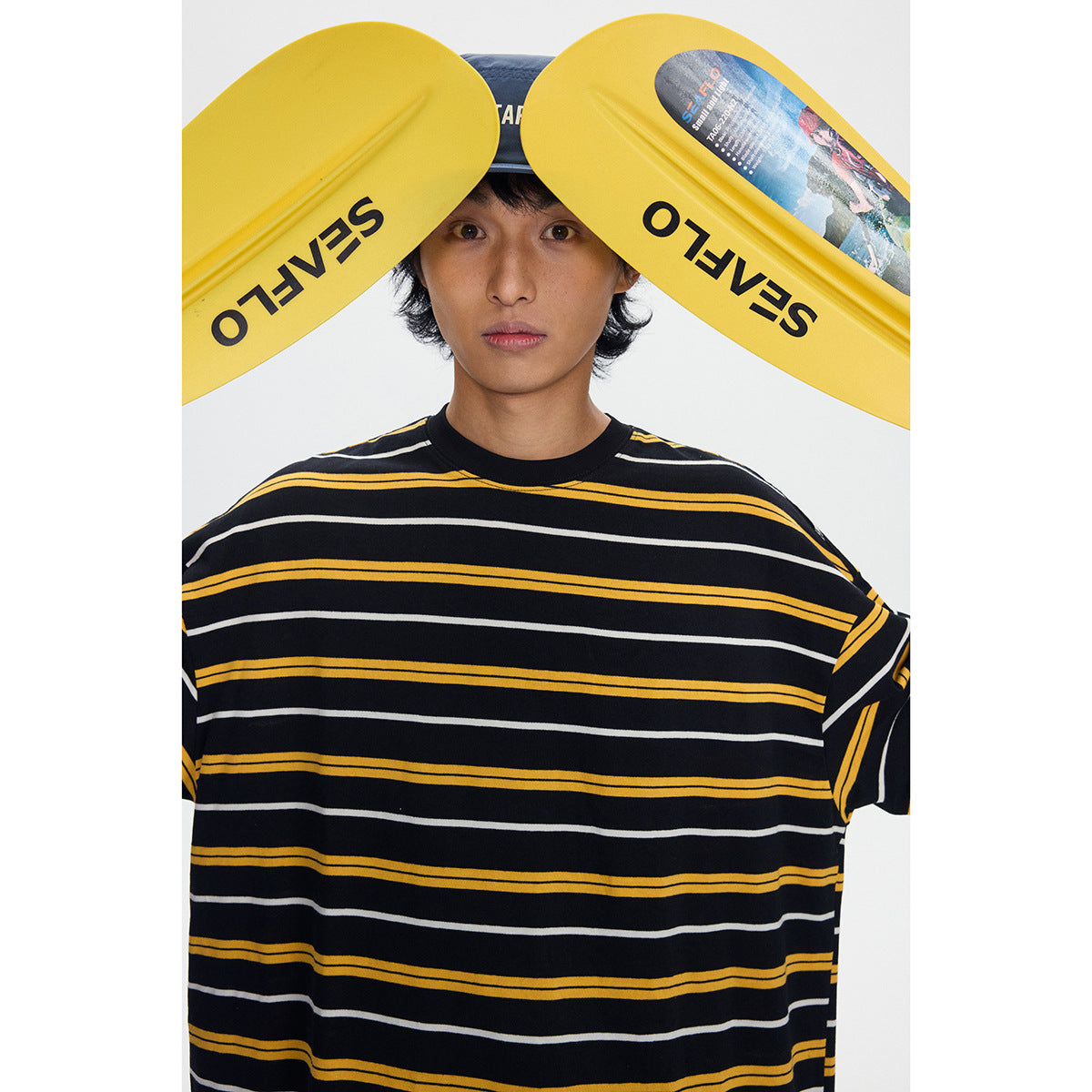 Close view of a model wearing a black oversized T-shirt with yellow and white horizontal stripes; yellow paddles frame his