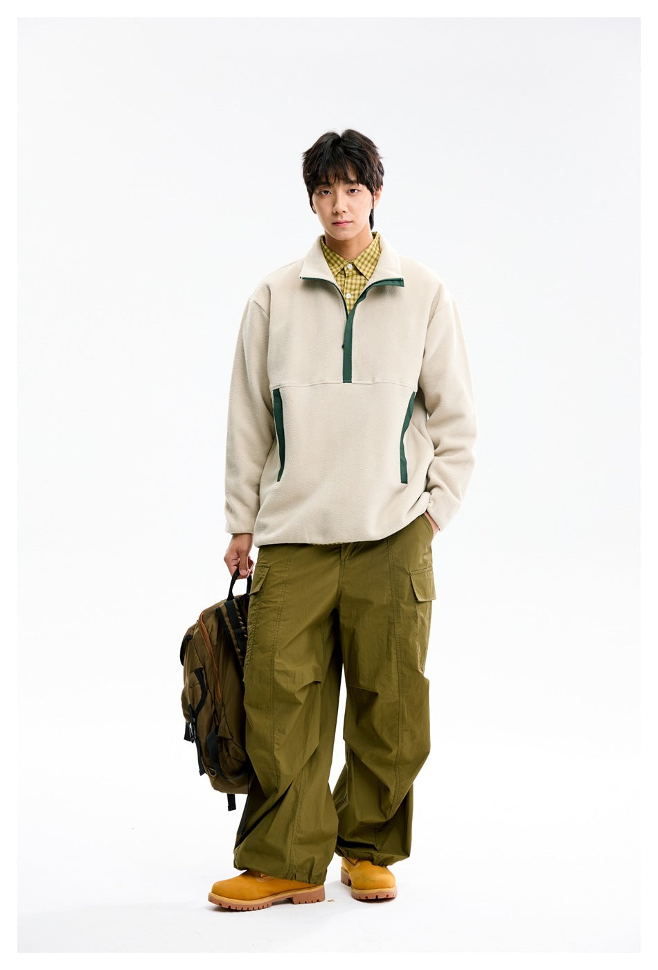 Model wearing beige half-zip fleece-style pullover with green trim, plaid shirt and olive cargo pants, holding a backpack