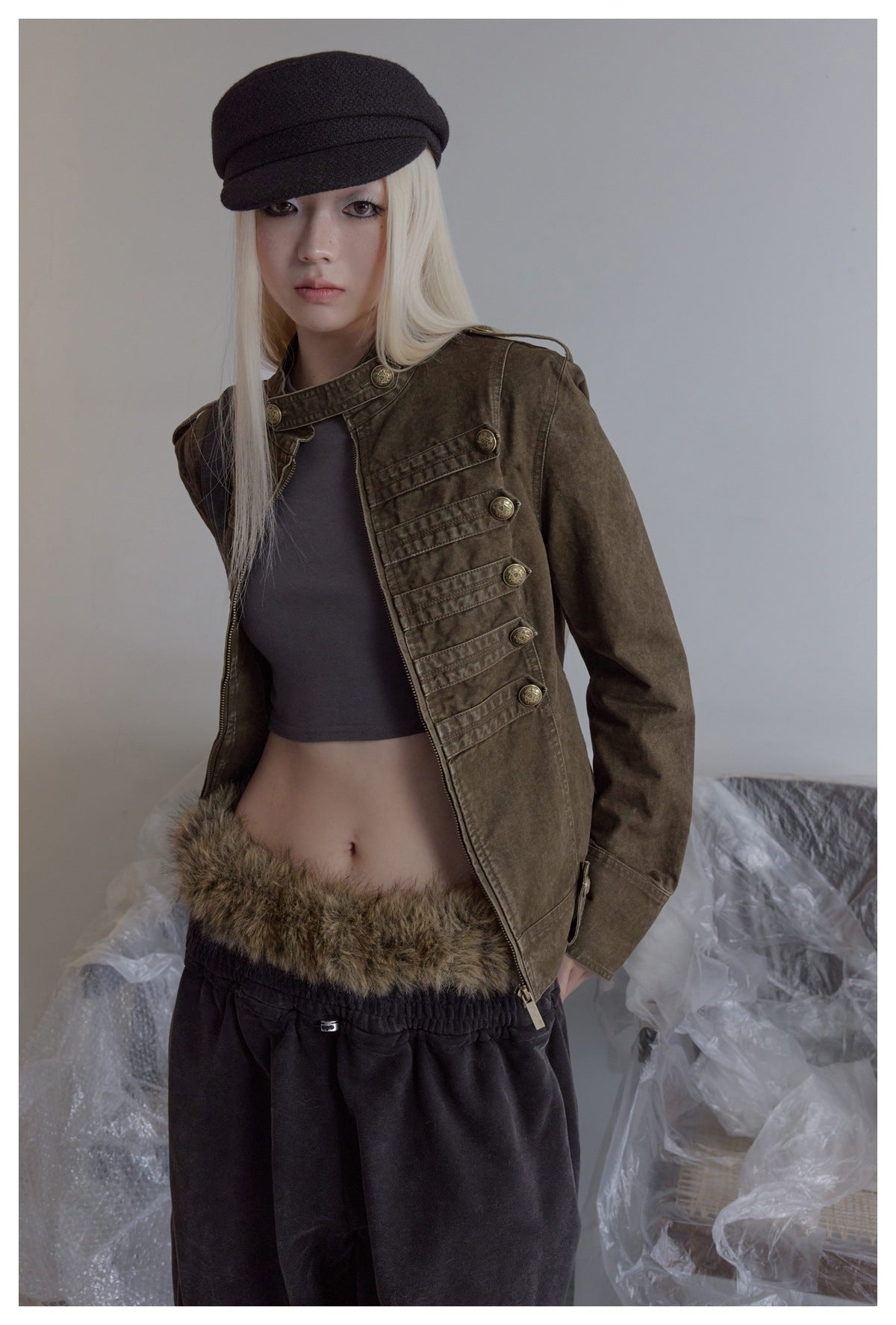Model wearing an open olive-brown military-style jacket with brass buttons over a dark cropped top.
