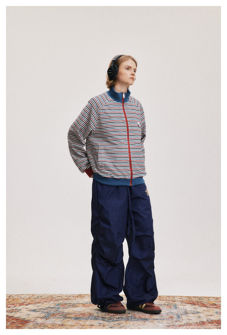 Model wearing a striped zip-up jacket with blue trim and loose navy pants, shown from the front at an angle.