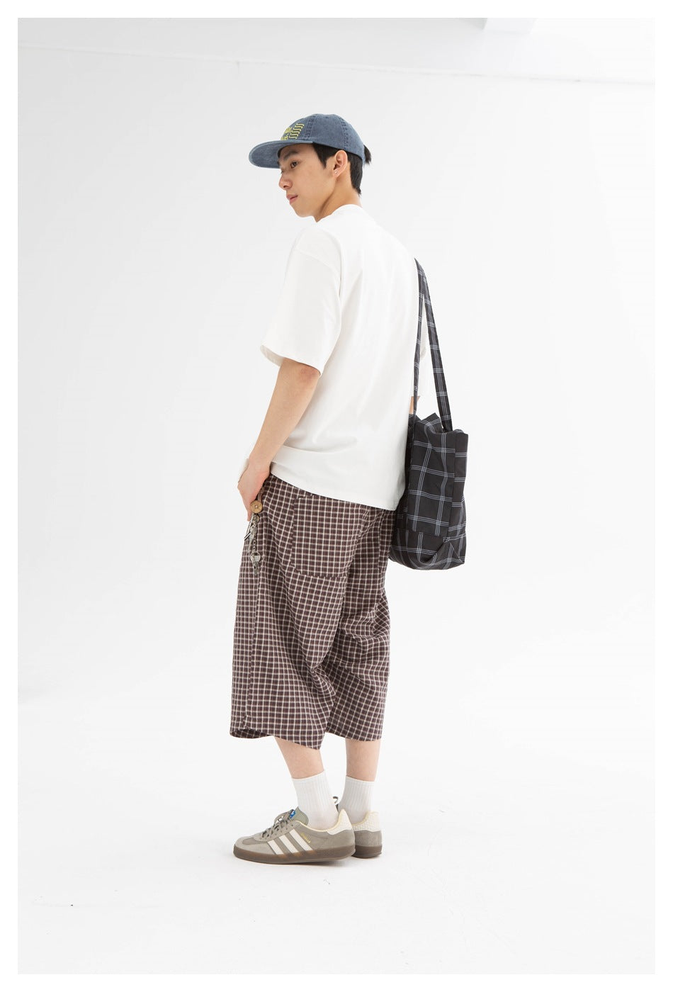 Model in a rear three-quarter view wearing brown plaid knee-length shorts, a white T-shirt and a black checked shoulder bag.