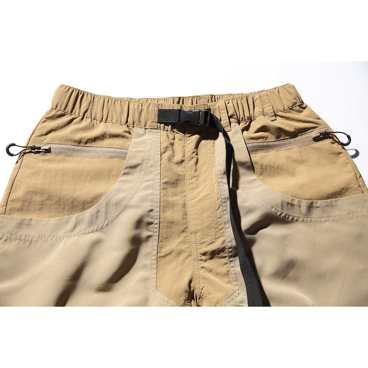 Close-up of tan shorts showing an elastic waistband, black webbing belt, and zippered front pockets.