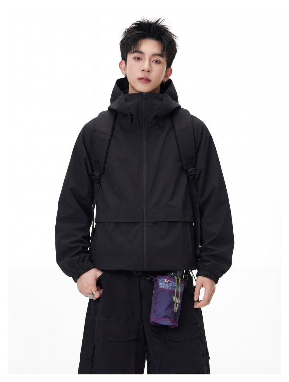 Model wearing a black hooded zip jacket with elastic cuffs and black cargo trousers, with backpack straps and a purple pouch.