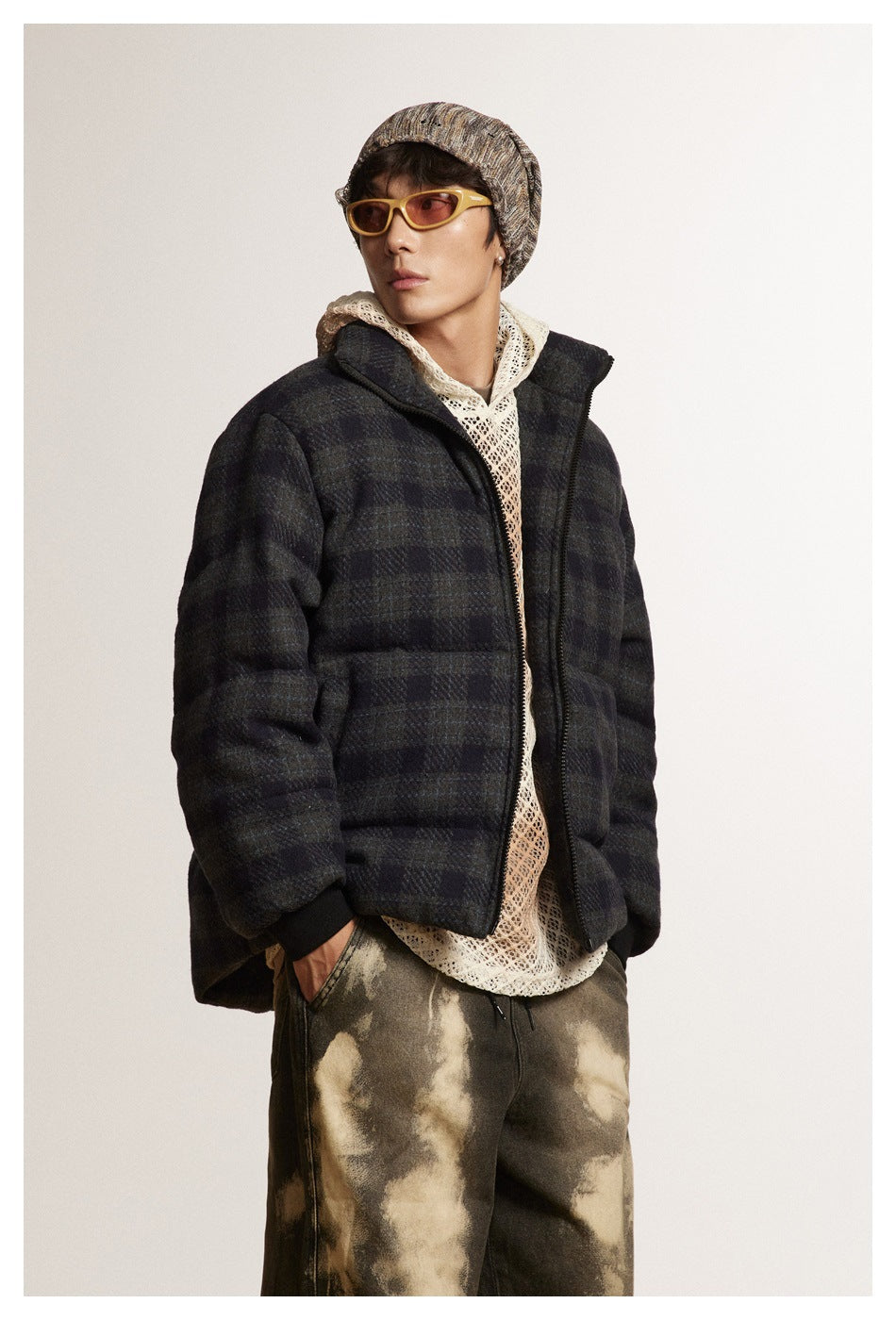 Model in a dark plaid puffer jacket over a cream open-knit hoodie, shown from the front with mottled brown pants.