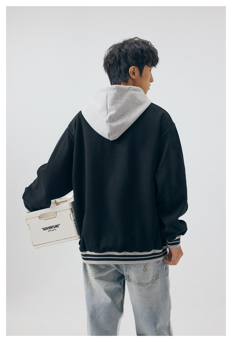 Rear view of a model wearing a black hoodie with a light gray hood and striped ribbed hem and cuffs.