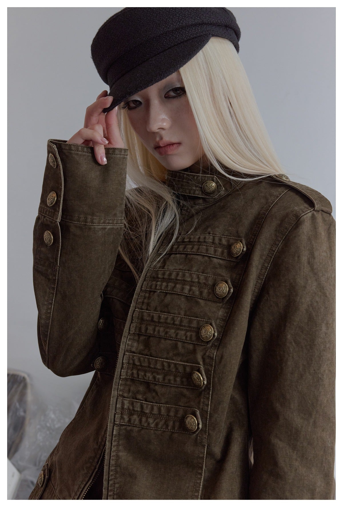 Model wearing a dark olive-brown military-style jacket with a high collar and rows of ornate buttons.
