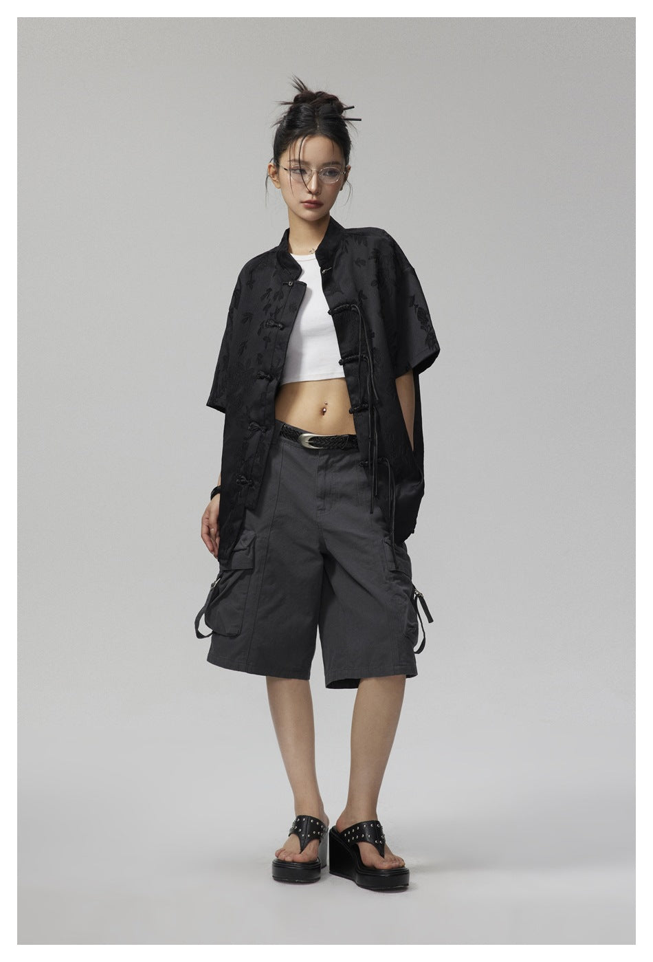 Full-length model wearing a black floral jacquard frog-button shirt over a white crop top and dark cargo shorts.