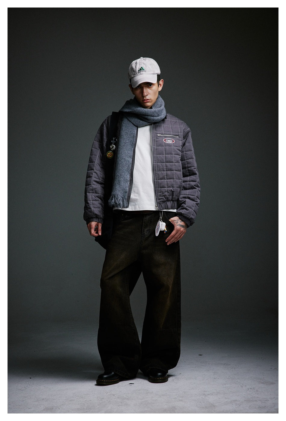 Full-length model wearing a gray quilted jacket, scarf, white shirt, dark wide-leg trousers and cap.