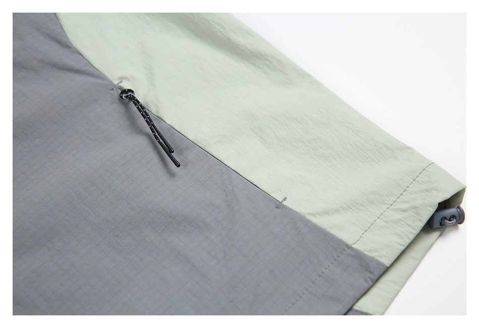 Close-up of a pale sage jacket sleeve with a gray panel, black zipper pull, and adjustable cuff tab.
