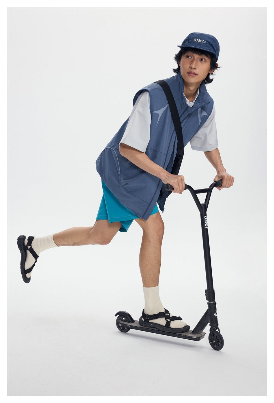 Model leaning on a scooter in a blue-gray patterned vest and turquoise shorts, with a white T-shirt and black bag.