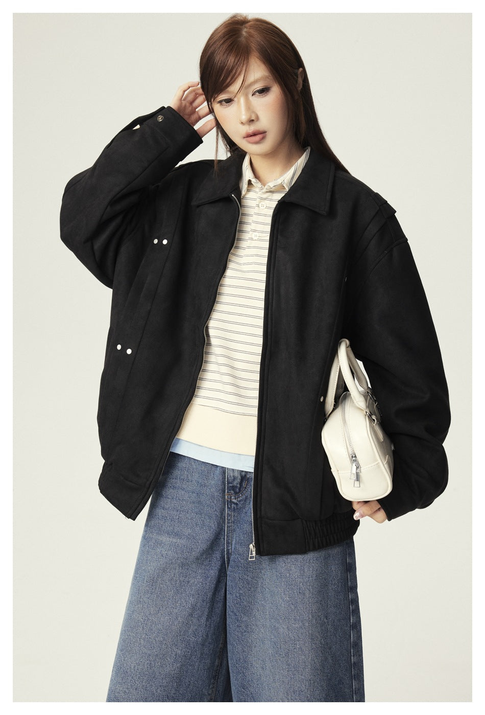 Model in an open black suede-look jacket with rivet details over a striped polo, holding a white handbag