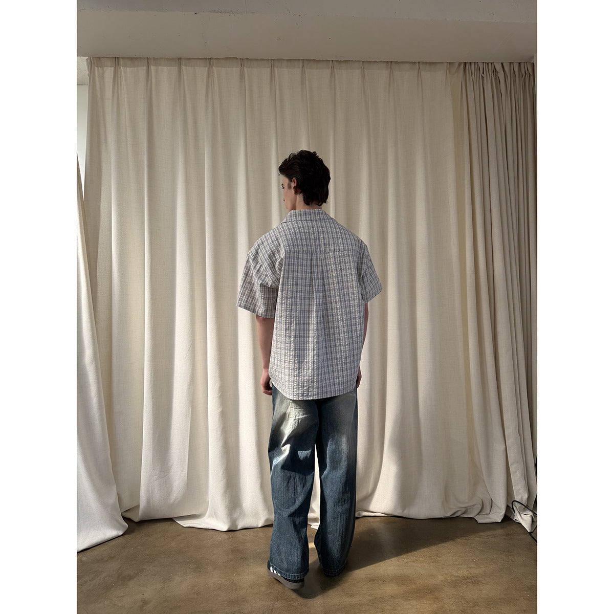 Back view of a man wearing a blue-gray plaid short-sleeve shirt with a center back pleat and loose dark jeans.