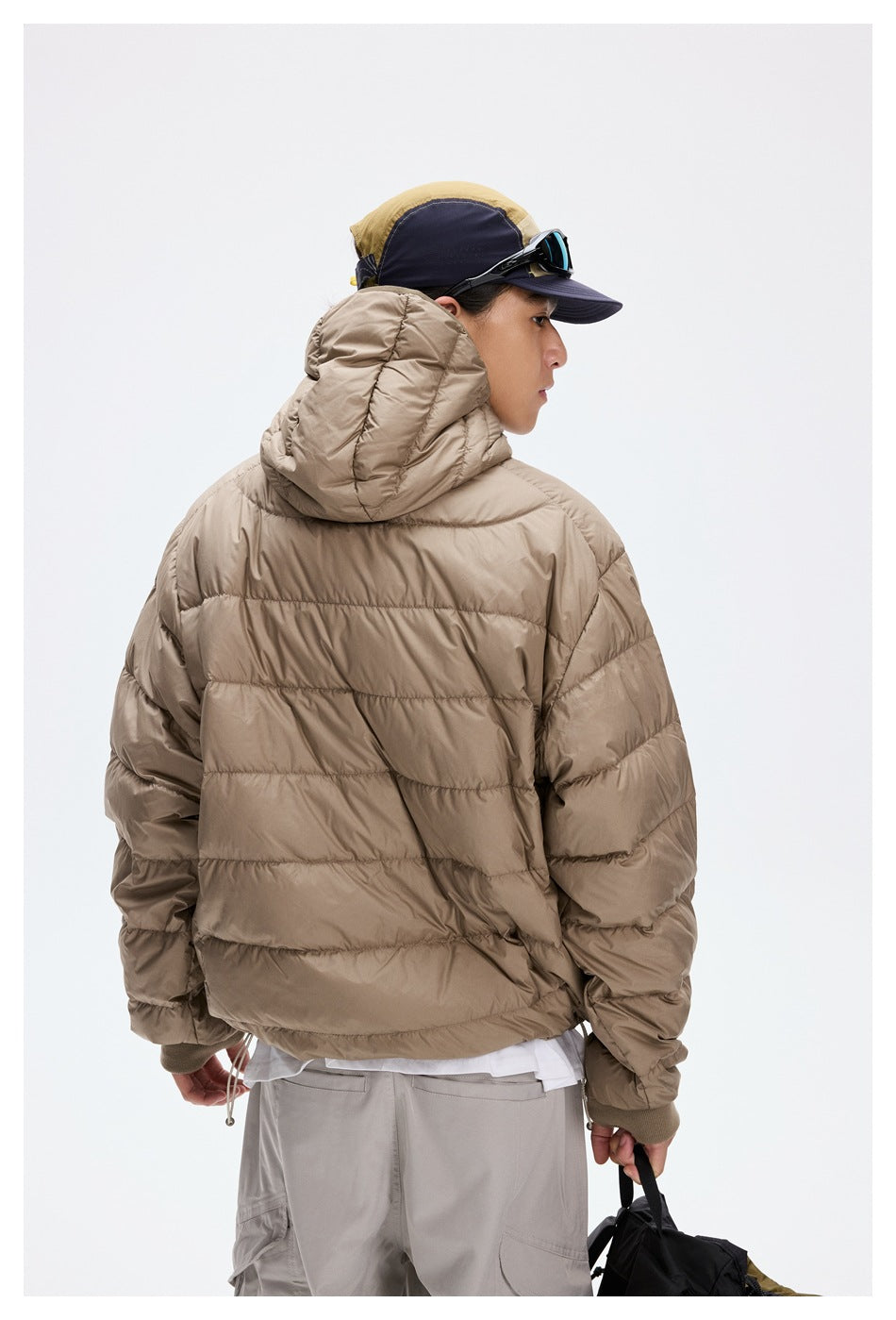 Back view of a model wearing a taupe hooded puffer jacket and light gray trousers.