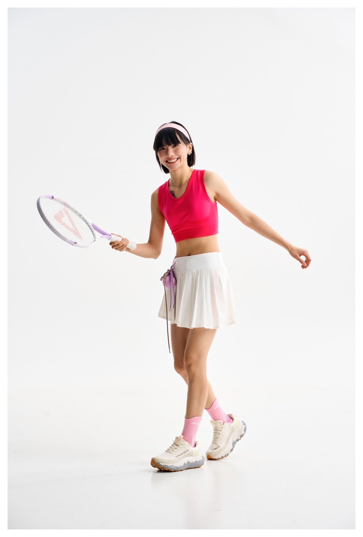 Model posing with a tennis racket in a bright pink crop top and white pleated tennis skirt.