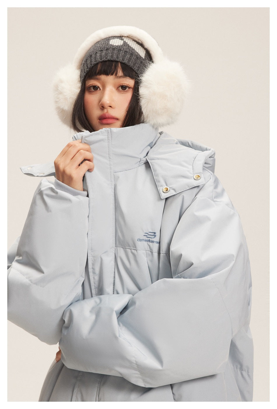 Close-up of a model in a light gray puffer jacket, showing the raised collar, hood details, snaps, and chest embroidery.