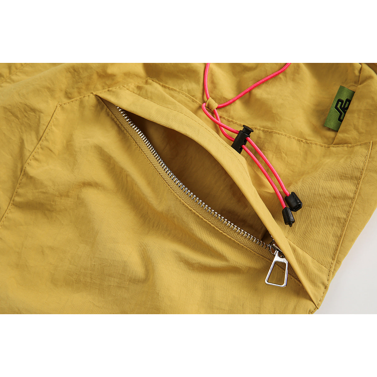 Close-up of mustard shorts showing a zip pocket, bright pink drawcord, and small green fabric tab.