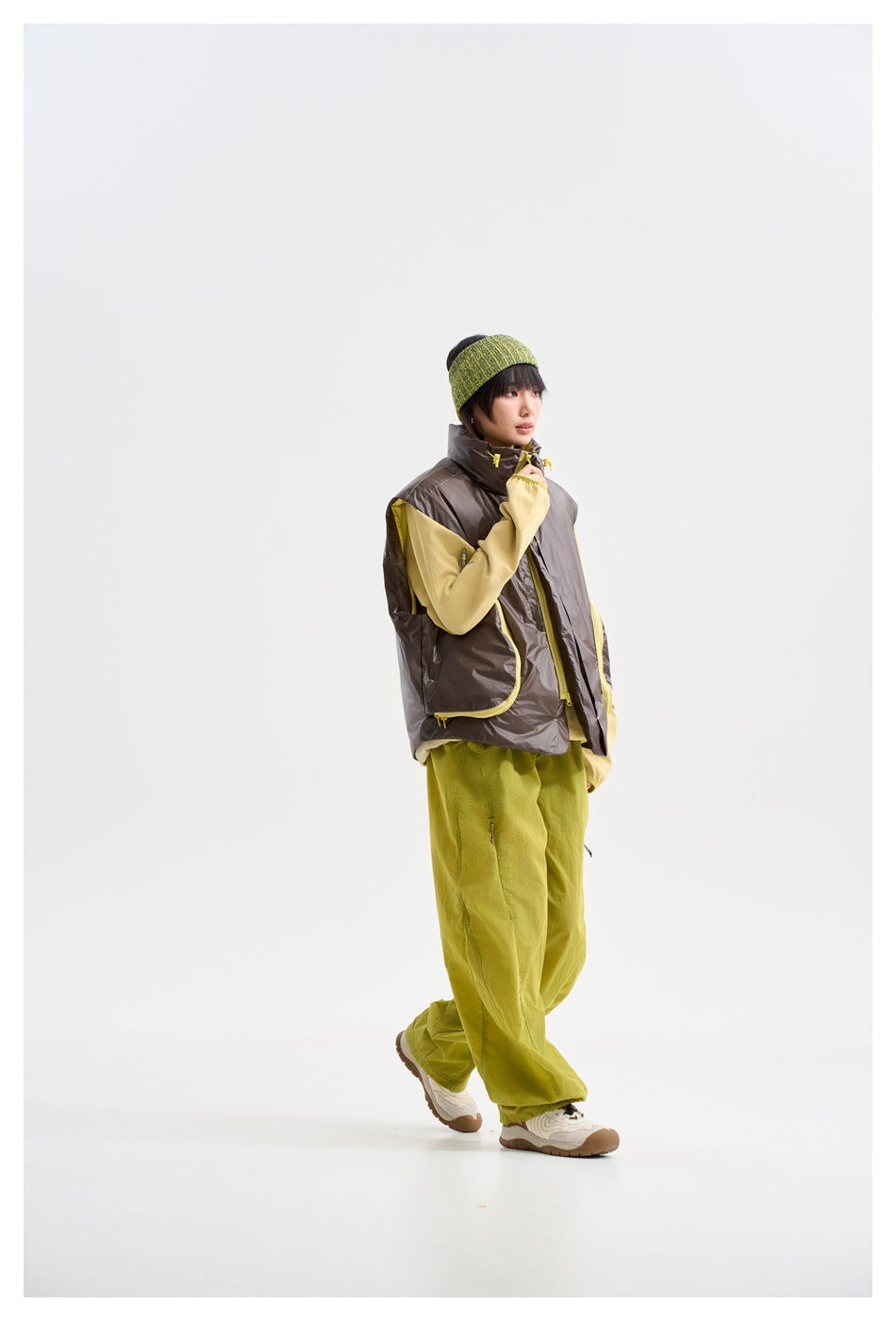 Three-quarter view of a model wearing a glossy charcoal vest with yellow-trimmed pockets over a pale yellow top and lime