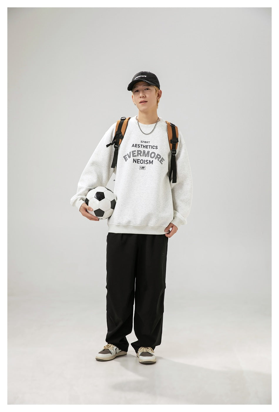 Model wearing a light gray graphic sweatshirt and black pants with a backpack, holding a soccer ball.
