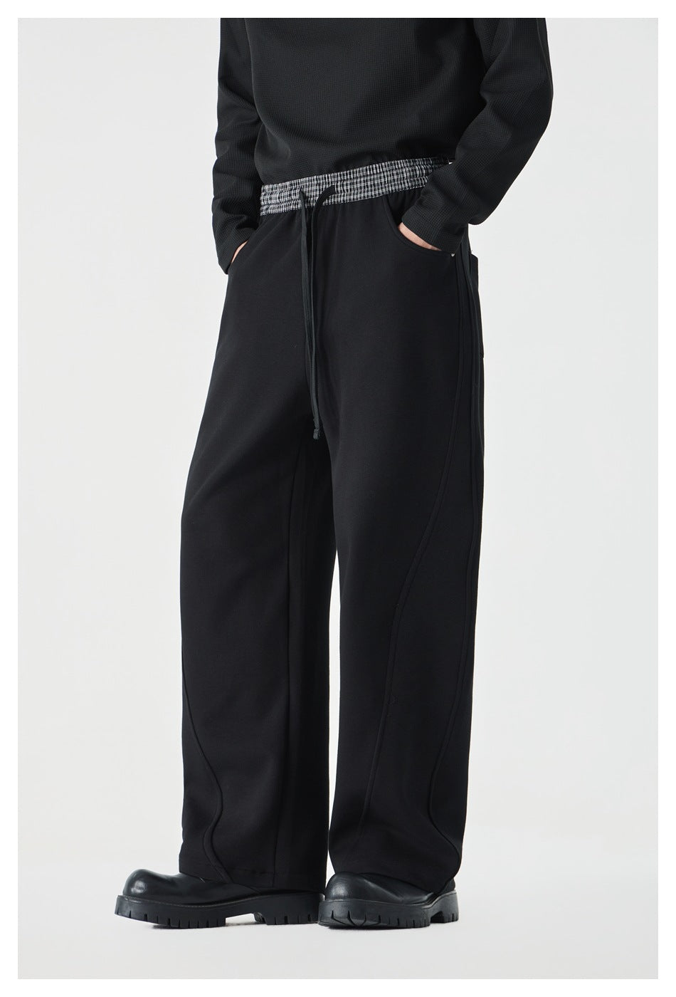 Model wearing black wide-leg pants with a checked waistband and curved seam details, hands in pockets.