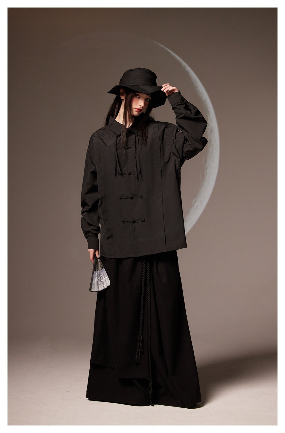 Model wearing a black collared shirt with frog closures, shown full length with a wide-brim hat.