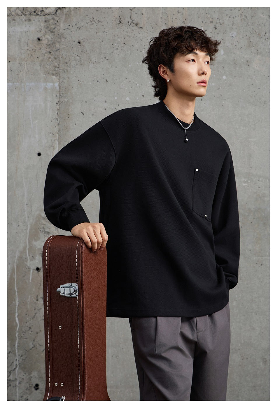 Model wearing a black oversized crewneck sweatshirt with a square chest pocket and loose gray trousers.