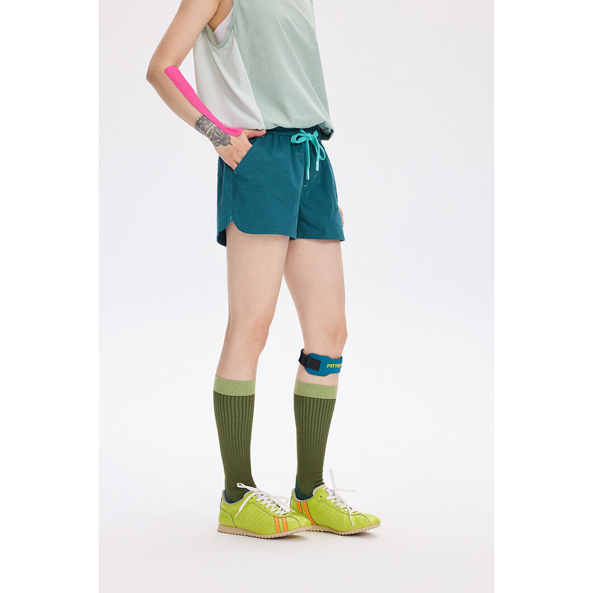 Model wearing dark teal drawstring shorts with a pale sleeveless top and knee-high socks; a pink mark overlays the arm.