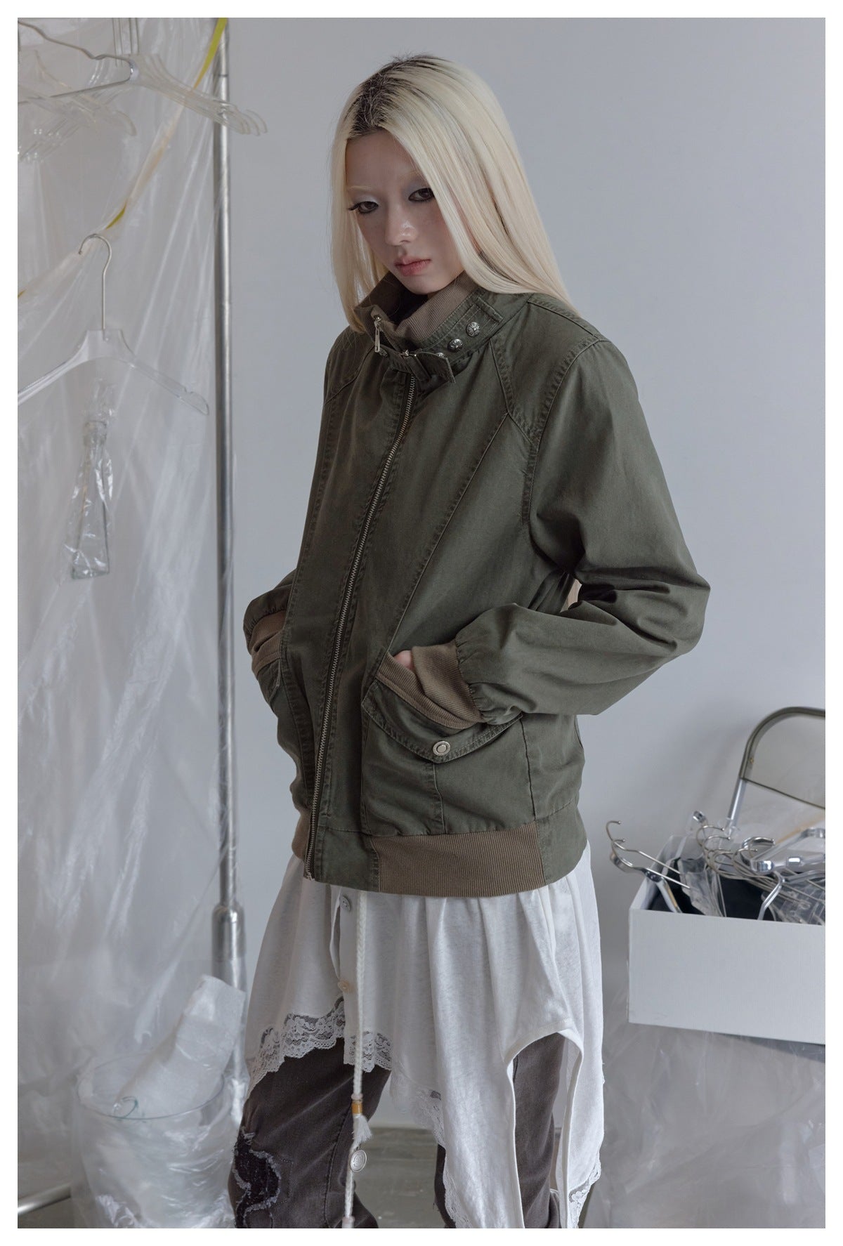 Model wearing an olive-gray zip jacket with a buckled stand collar, brown ribbed cuffs and hem, and flap pockets.