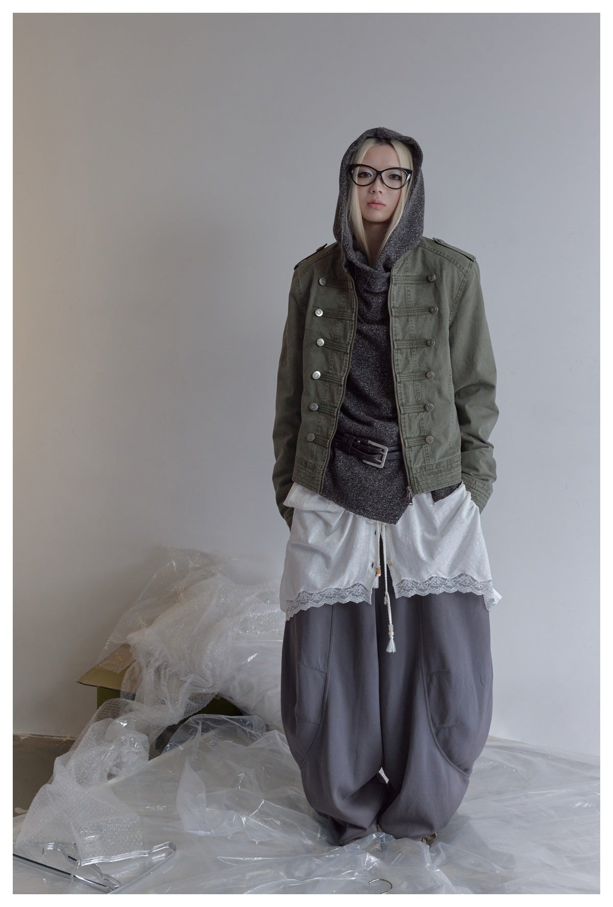 Model wearing an olive button-detailed jacket over a charcoal hooded top, white lace-trim layer, and loose gray trousers.