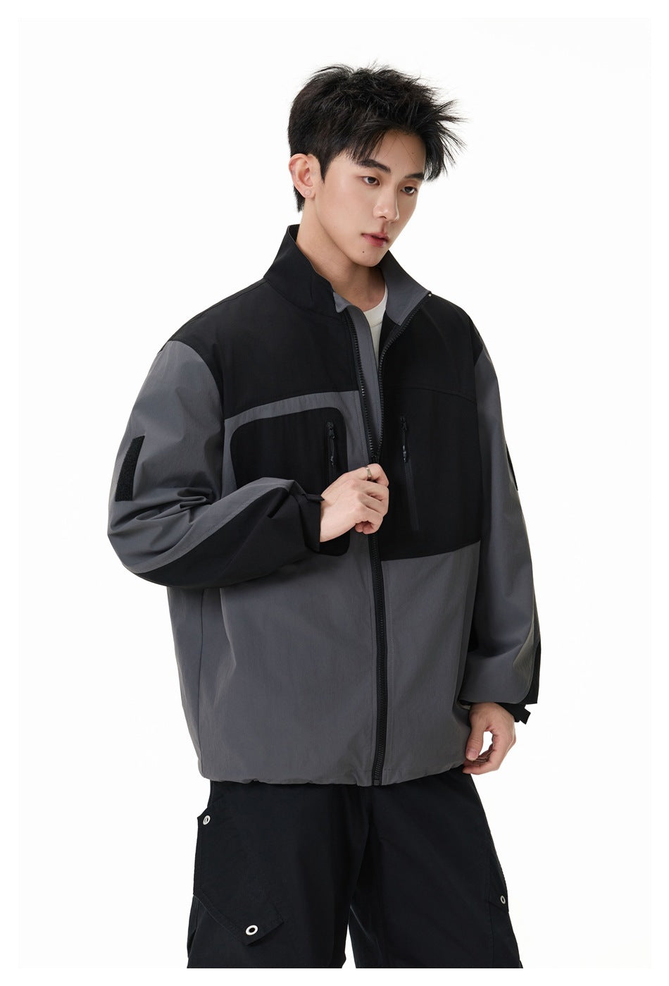 Model wearing a black and charcoal-gray color-block jacket with a high collar and two vertical zip chest pockets.
