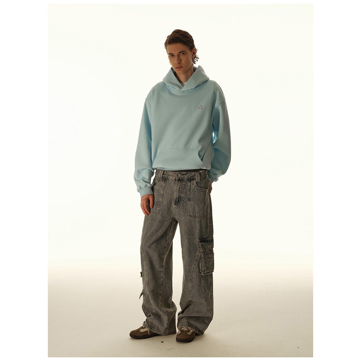Model wearing a pale blue hooded sweatshirt with a small chest emblem and loose gray cargo jeans.