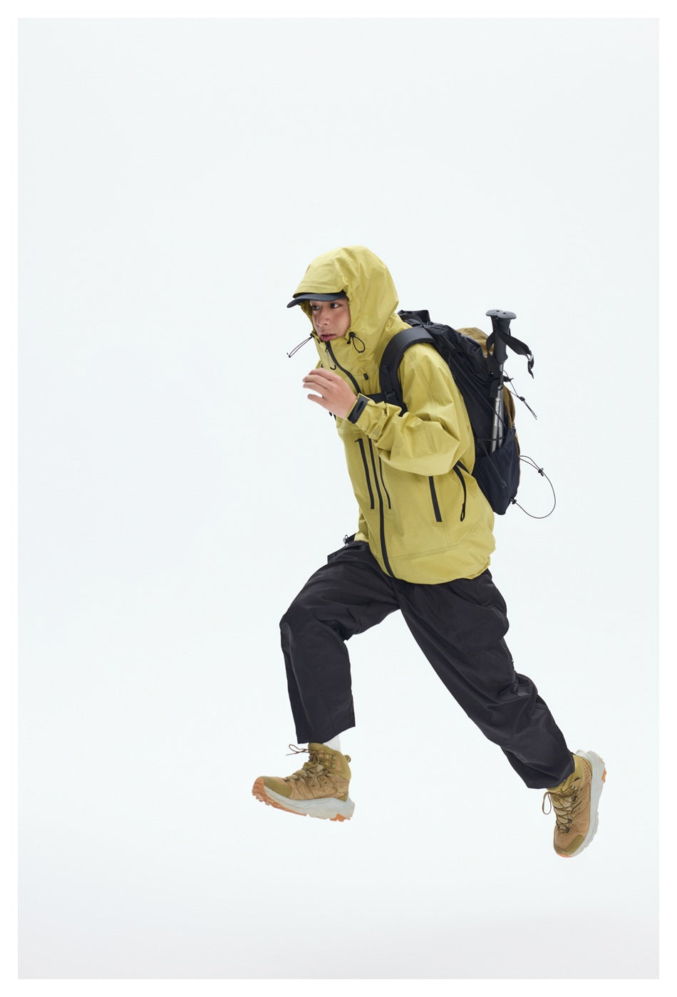Hiker in a yellow hooded jacket and black pants strides with a loaded backpack and trekking poles.