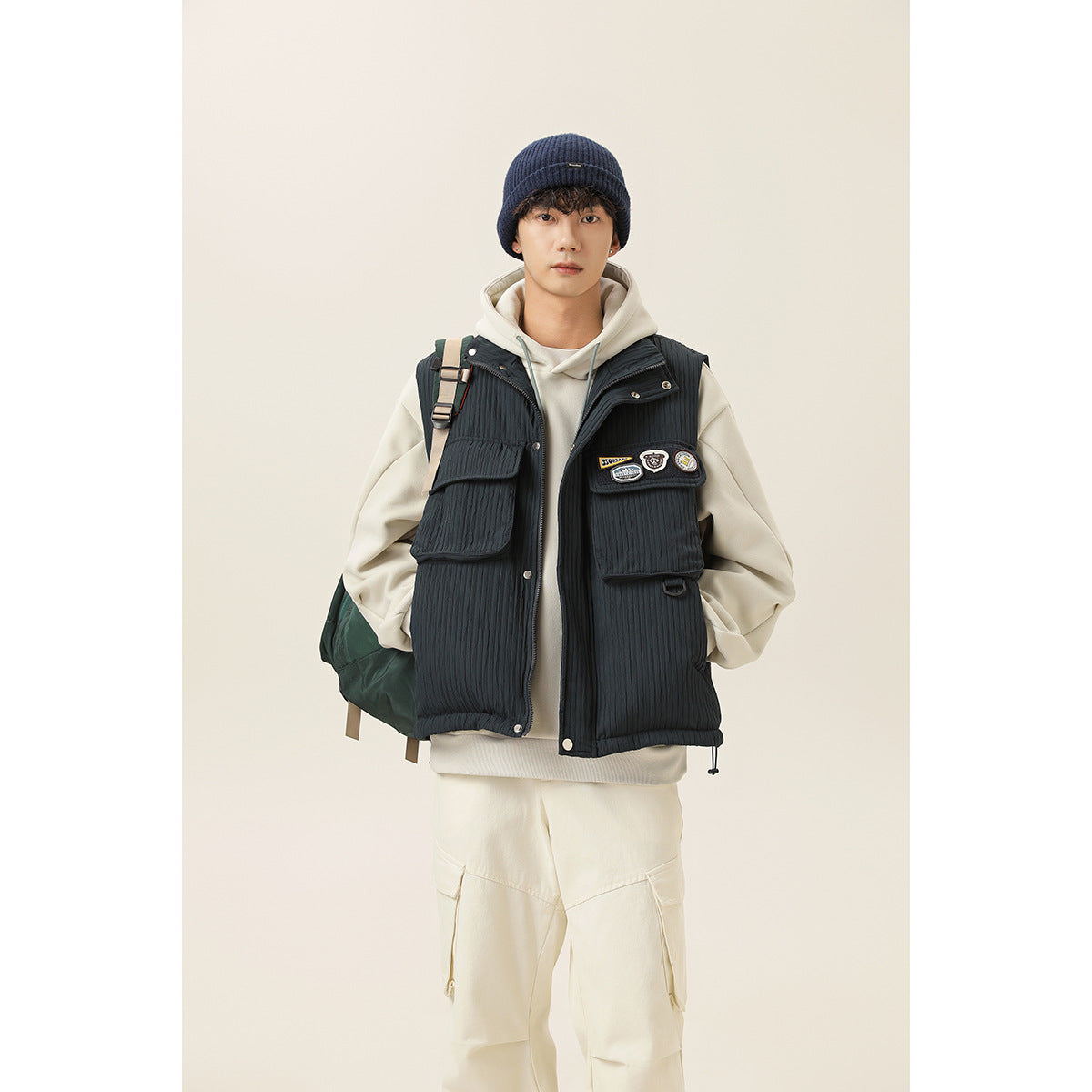 Model wears a dark navy utility vest with patch details over a cream hoodie and matching cargo pants.