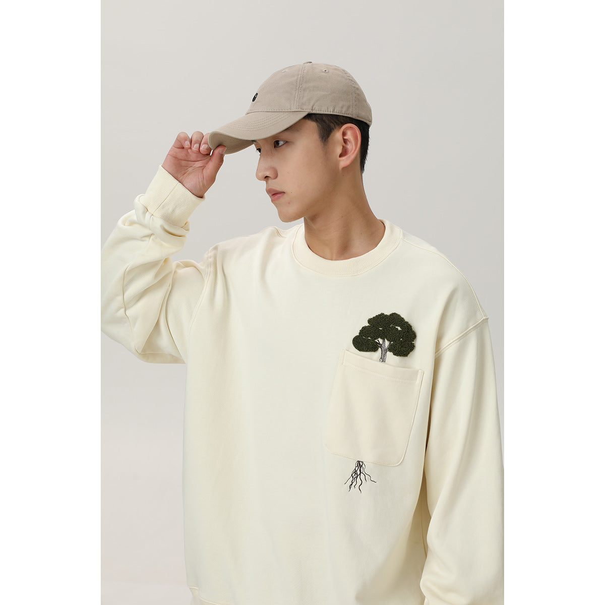 Model wearing an ivory crewneck sweatshirt with a chest pocket and embroidered tree-and-roots design.