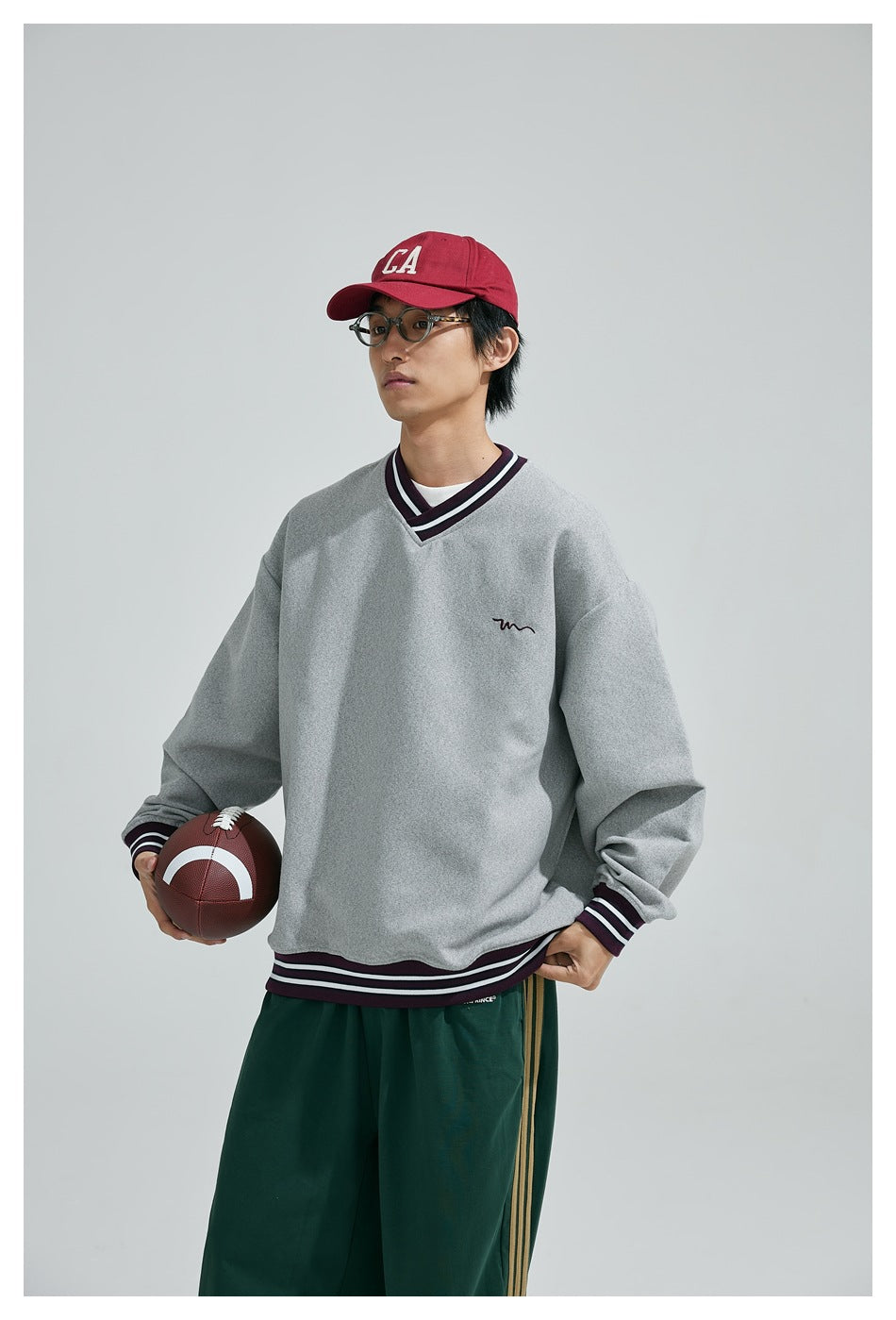 Model wearing a light gray V-neck sweatshirt with dark striped trim and green track pants, holding a football.