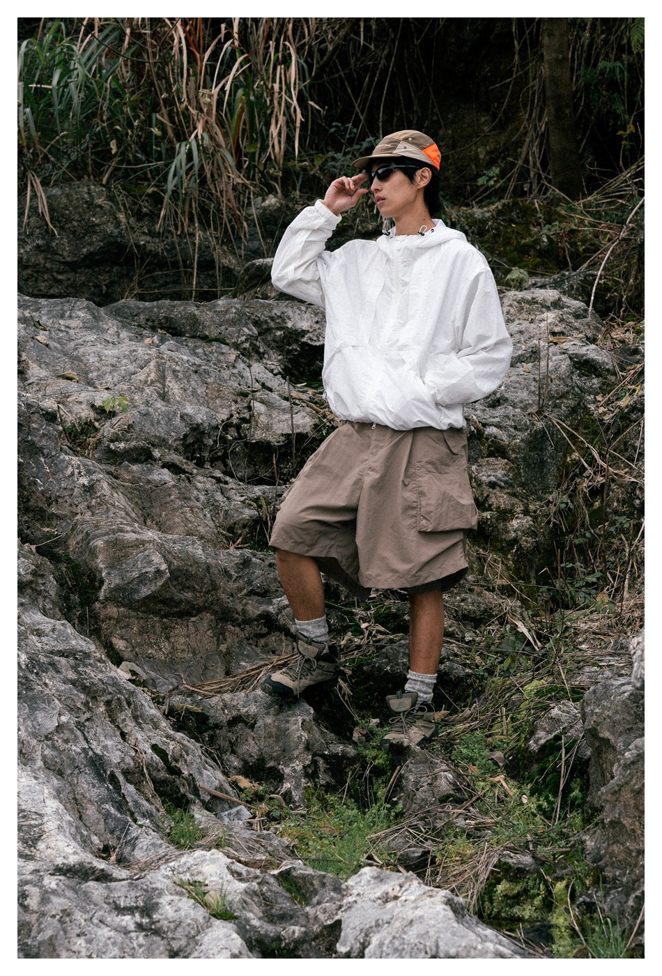 A man stands among rocks wearing a white hooded pullover and loose taupe cargo shorts with large side pockets.