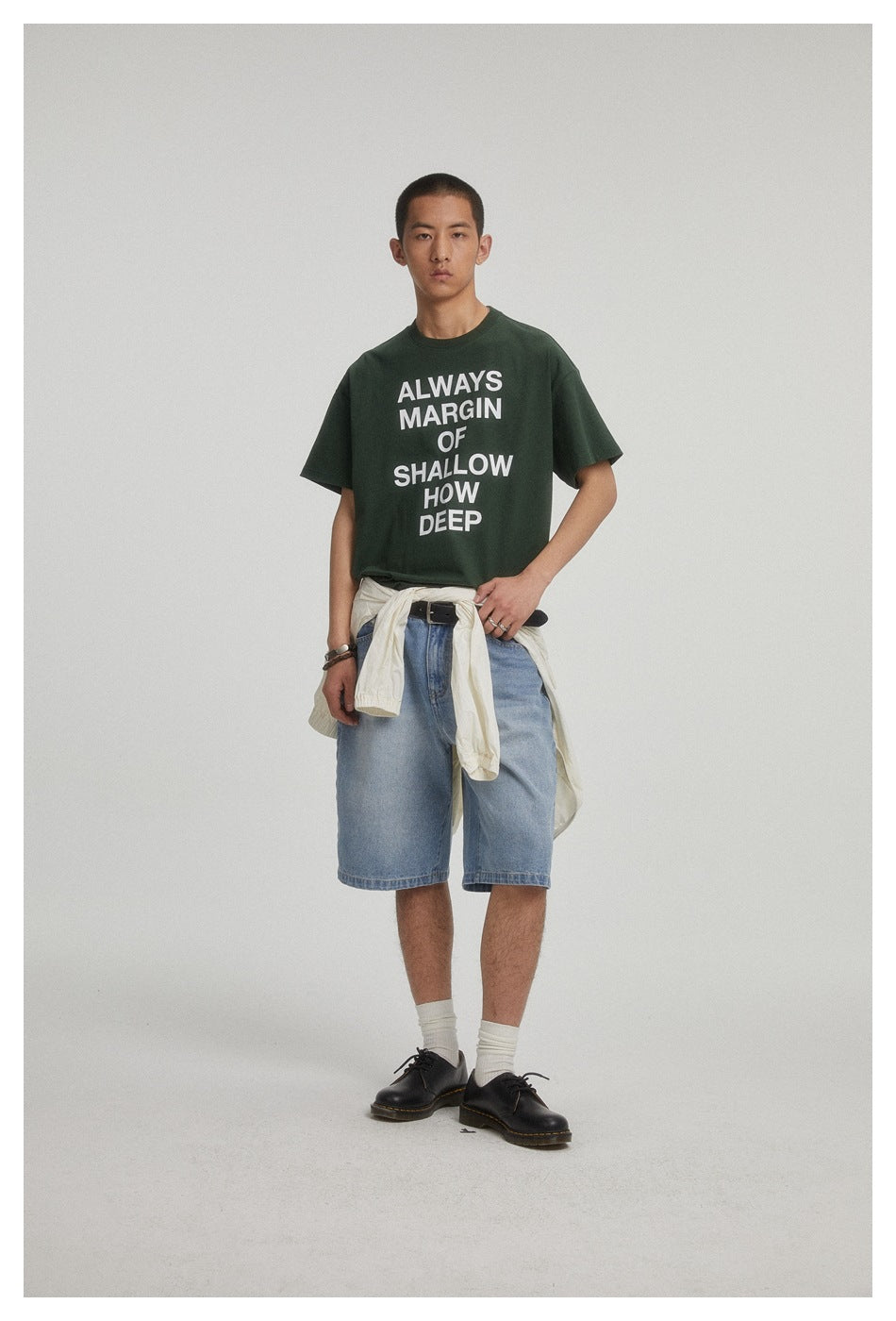 Model wearing a dark green text-print T-shirt and light blue denim shorts, with a light shirt tied at the waist.