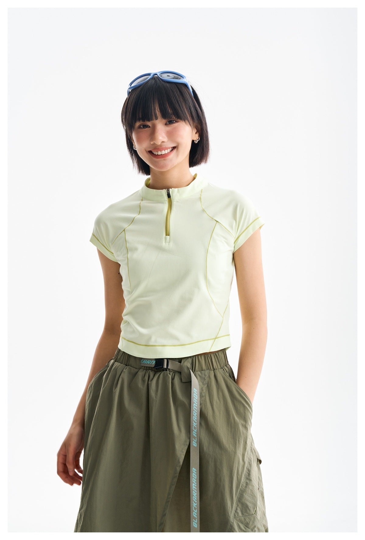 Model wearing a pale yellow short-sleeve zip-neck top with curved contrast seams and an olive skirt.