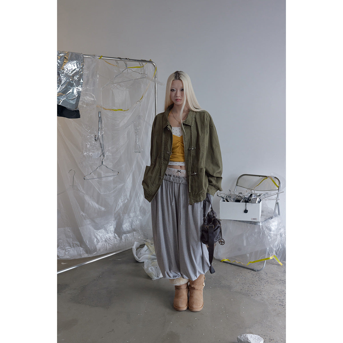 Model wearing an oversized olive jacket with silver hook closures and a gathered collar, paired with gray wide-leg pants.