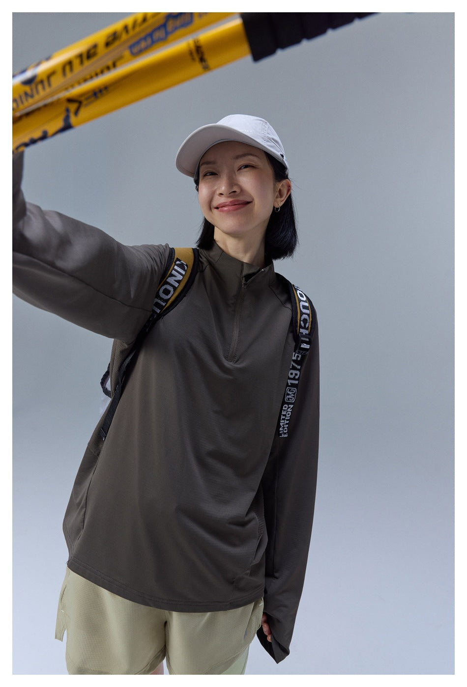 Close view of a model wearing a dark taupe quarter-zip top and khaki shorts, with hiking poles crossing the frame.