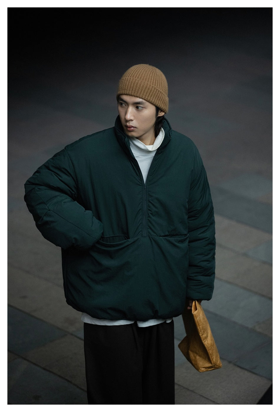 Man wearing a dark green half-zip padded jacket with a tan beanie, white turtleneck and dark trousers, holding a paper bag